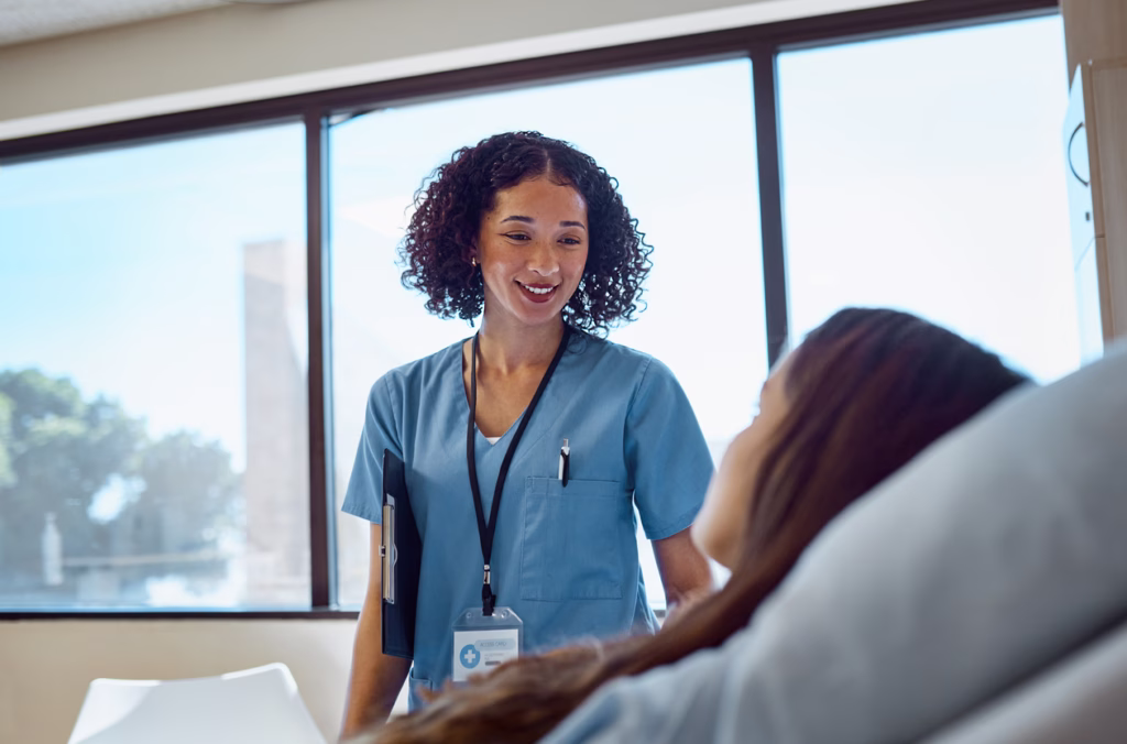 Female nurse with curly hair, smiling at patient lying in bed beside window